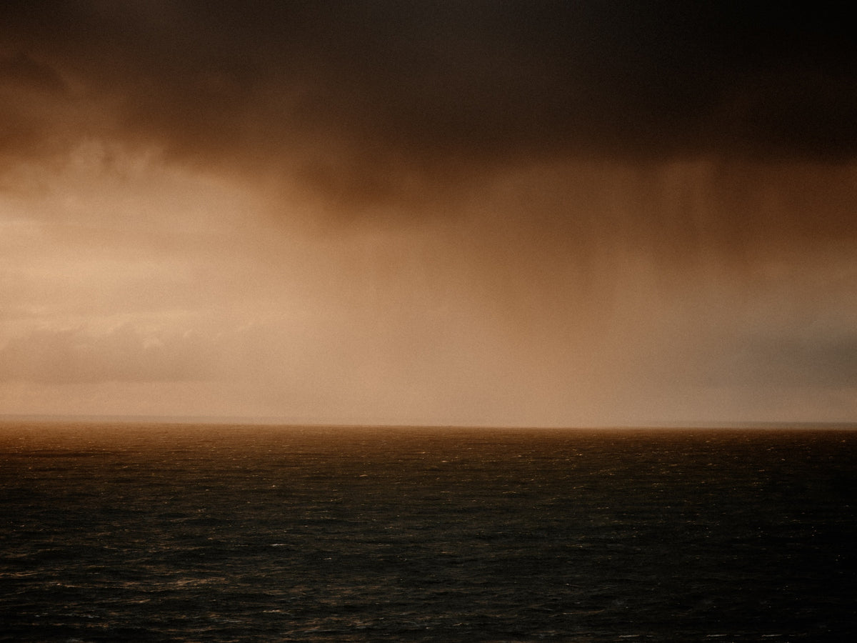 A dramatic seascape with dark storm clouds over a calm ocean, lit by warm golden light near the horizon.