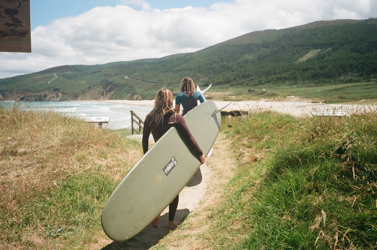 Two people wearing wet suits carry surf boards down a beach path to the sea