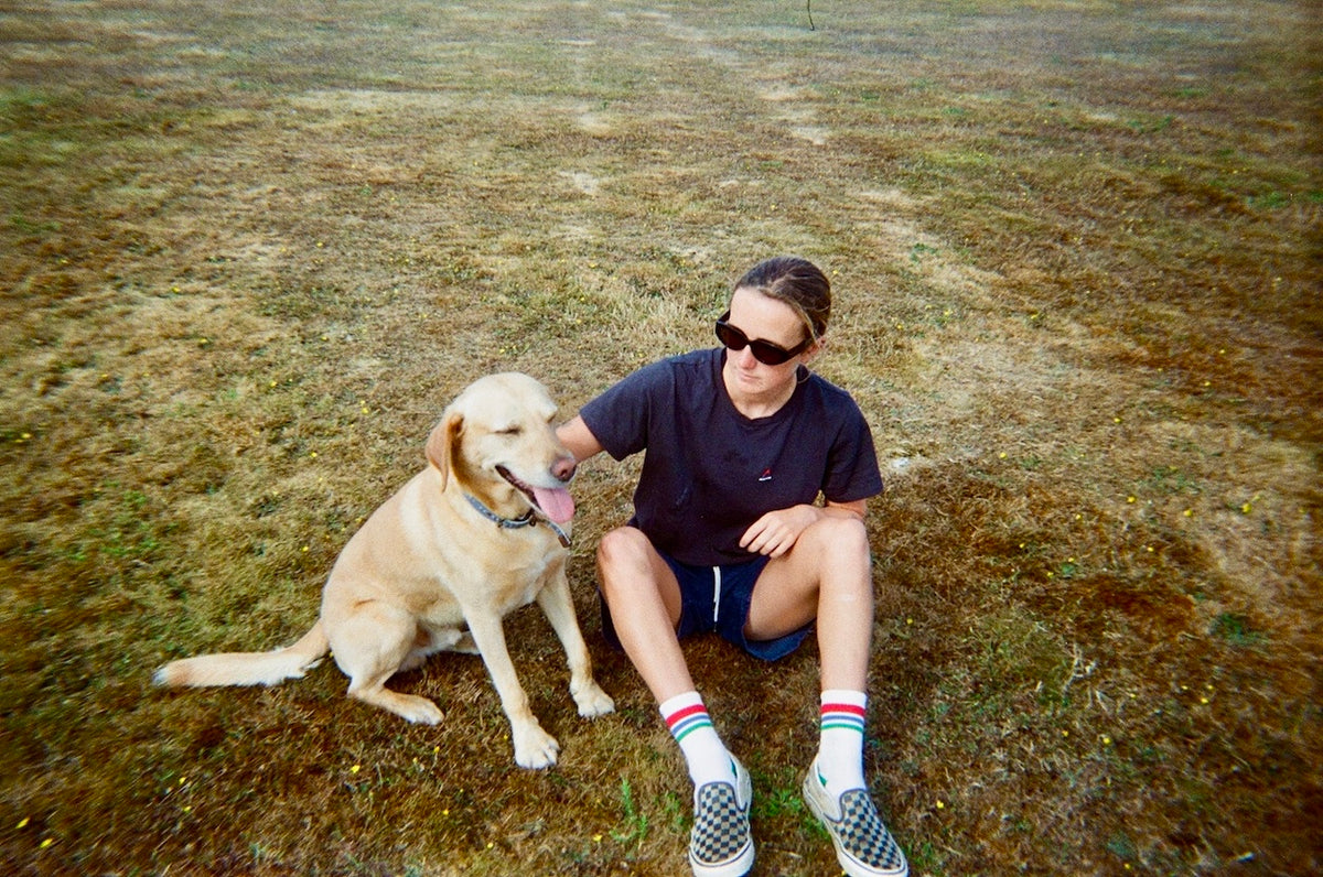 A woman wearing a black t-shirt, shorts, and slip-on shoes sits on dry grass next to a light yellow Labrador retriever dog.