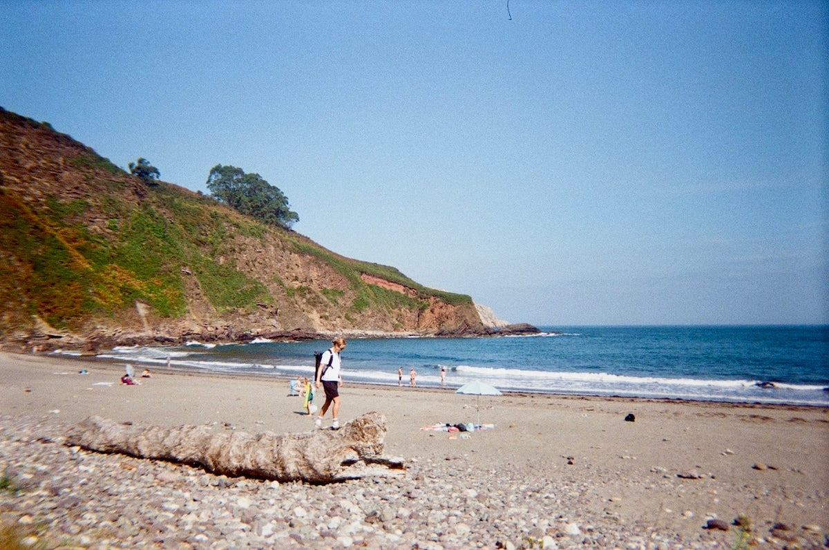 A scenic beach landscape with a sandy shore, pebbled foreground, blue ocean, and a grassy rocky cliff under a clear sky. A few small people are visible near the water and on the beach.