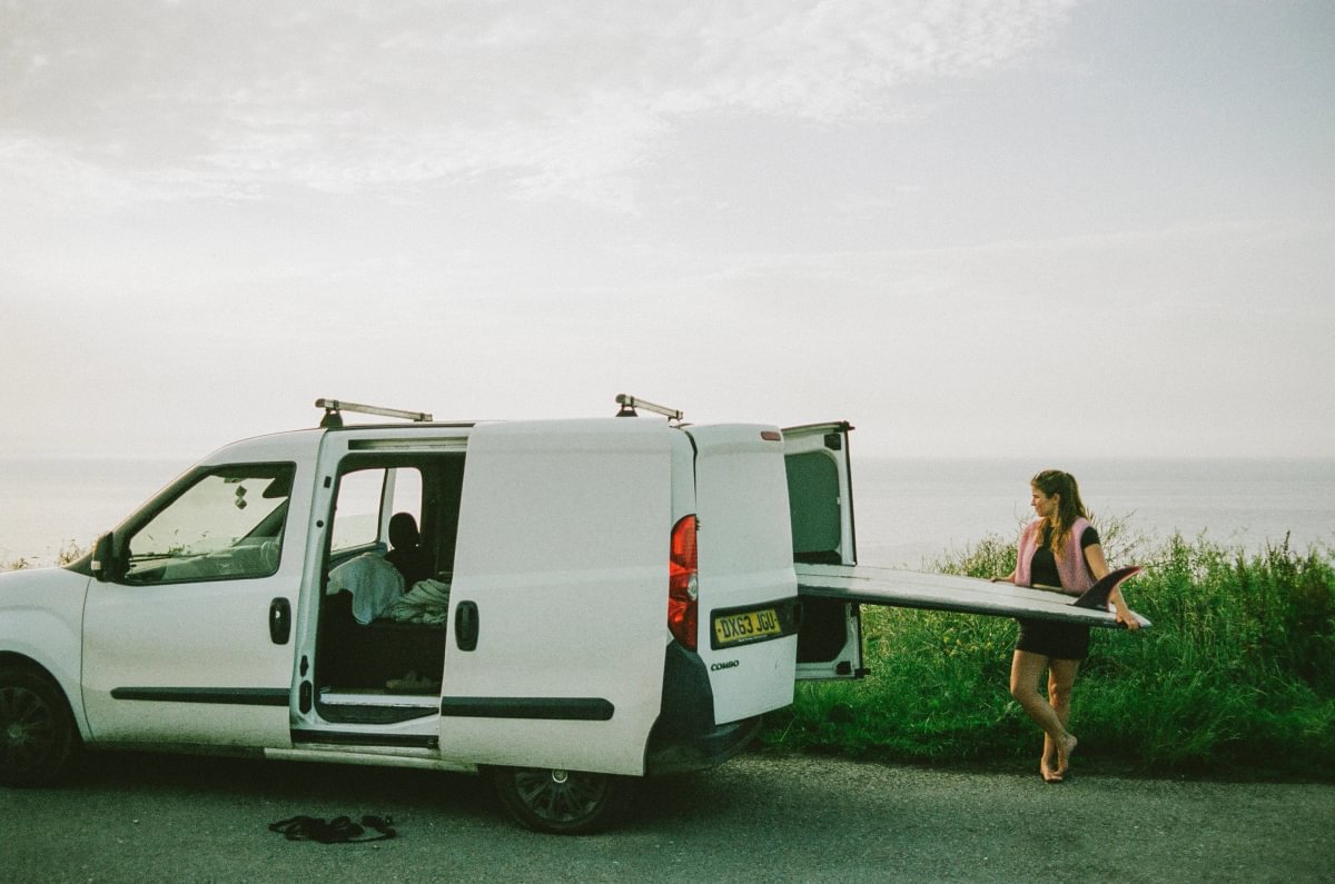 A white van parked outdoors by the sea with its side and rear doors open, and a woman carrying a surfboard beside the vehicle.